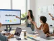 Tim kreatif mendiskusikan GEGESLOT di kantor modern dengan laptop dan papan tulis.
